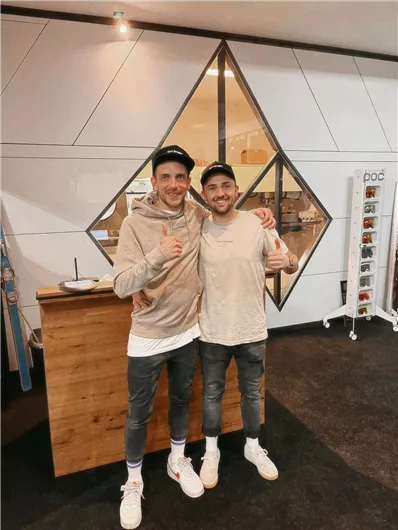 Zwei lächelnde Männer stehen in einem modernen Innenraum und geben Daumen hoch. Im Hintergrund ist eine stilvolle Glaswand zu sehen.