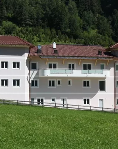 Ein modernes Gebäude umgeben von grünen Wiesen und Bergen. Das Haus hat eine helle Fassade und einen roten Dachstuhl.