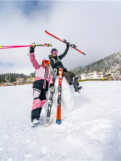 Zwei Skifahrerinnen stehen im Schnee und feiern. Im Hintergrund sind verschneite Berge und Hütten zu sehen.