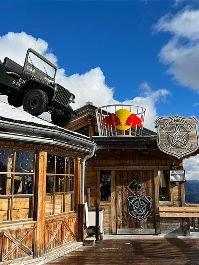 Ein einzigartiges Holzgebäude in den Bergen mit einem Jeep auf dem Dach. Es ist eine verschneite Landschaft und der Himmel ist teilweise bewölkt.