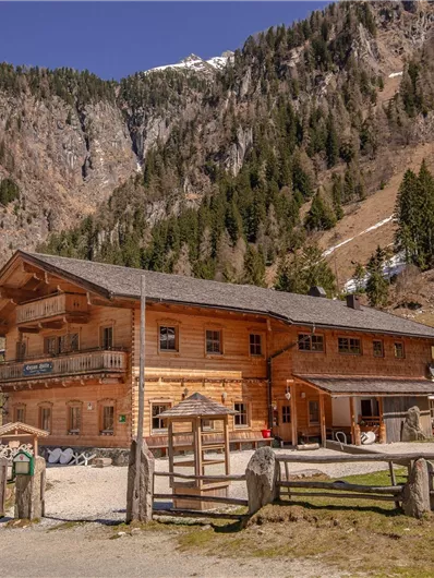 Ein traditionelles Holzhaus vor einer beeindruckenden Berglandschaft. Die Umgebung zeigt grüne Wiesen und schneebedeckte Gipfel.