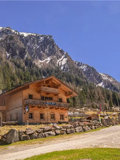 Ein altes Holzhaus in den Bergen, umgeben von einer malerischen Landschaft. Im Hintergrund sind schneebedeckte Gipfel und grüne Wiesen zu sehen.