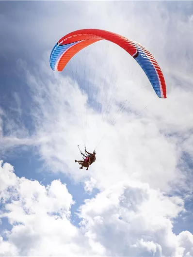 Ein Gleitschirmflieger schwebt durch den blauen Himmel mit weißen, wolkigen Elementen. Der Gleitschirm hat rot-blaues Design.