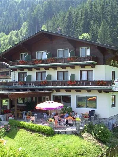 Ein malerisches Gästehaus in den Bergen, umgeben von grüner Natur. Auf der Terrasse sind Tische und Stühle mit Sonnenschirmen.