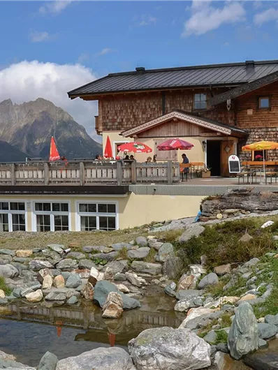 A cozy mountain house with red sun umbrellas. In the background, the mountains and a clear sky are visible.
