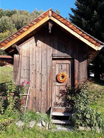 A rustic wooden house with a traditional roof. An orange wreath hangs in front of the door, surrounded by colorful plants.