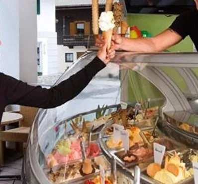 Een vrouw geniet van een ijsje terwijl ze vriendelijk met een verkoper praat. Op de achtergrond is een selectie van verschillende ijssoorten zichtbaar.