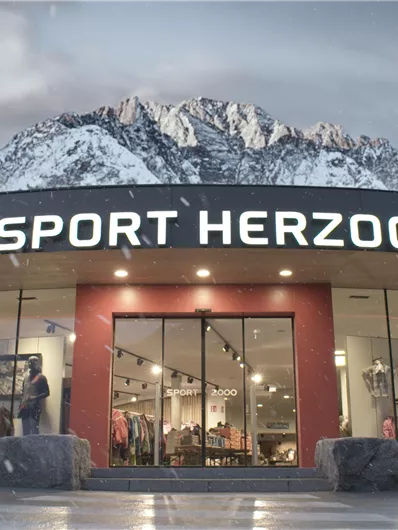 Ein sportliches Geschäft namens "Sport Herzog", umgeben von schneebedeckten Bergen. Die Fassade ist modern und einladend.
