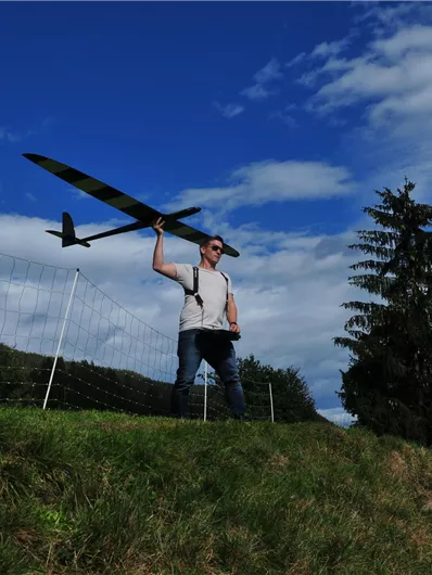 Ein Mann steht auf einem Hügel und hält ein Modellflugzeug in der Hand. Im Hintergrund sind ein blauer Himmel und einige Bäume zu sehen.