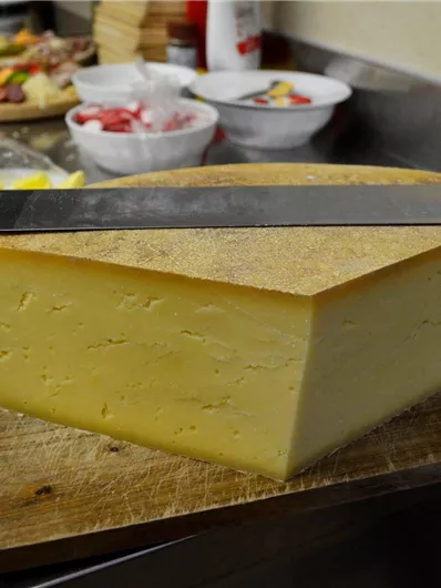 Een groot stuk kaas ligt op een houten snijplank, naast een groot mes. Op de achtergrond zijn meer voedingsmiddelen in schalen te zien.