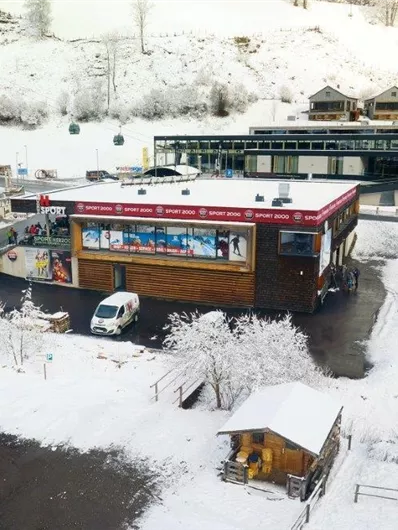 Ein schneebedecktes Dorf mit einem modernen Gebäude und mehreren Skihütten. Autos stehen auf dem Parkplatz, umgeben von einer winterlichen Landschaft.