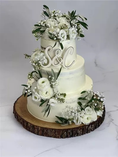 Een elegante drie-laagse bruidstaart met witte bloemen en groene bladeren. De taart is geplaatst op een houten plank en heeft de initialen "S&C" gedecoreerd.