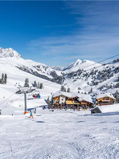 Een schilderachtig sneeuwlandschap met skiërs en een gezellig berghuis. Op de achtergrond zijn besneeuwde bergen en een blauwe lucht te zien.