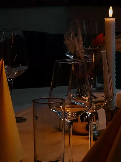 An elegantly set table with glasses and yellow napkins. A candle burns in the center, providing atmospheric light.