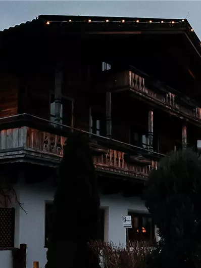 An old wooden house with a balcony and warm lighting. The sky is overcast and the surroundings seem calm.