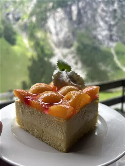 Een stuk fruitige cake met perziken en een roomtopping. Op de achtergrond zijn groene bergen te zien.