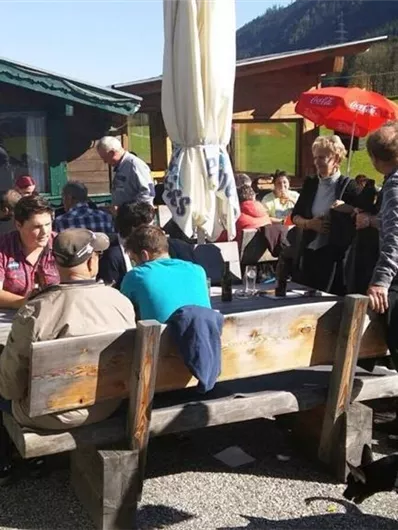 A lively terrace with wooden benches and tables. People are enjoying time outside in a beautiful setting with mountains in the background.