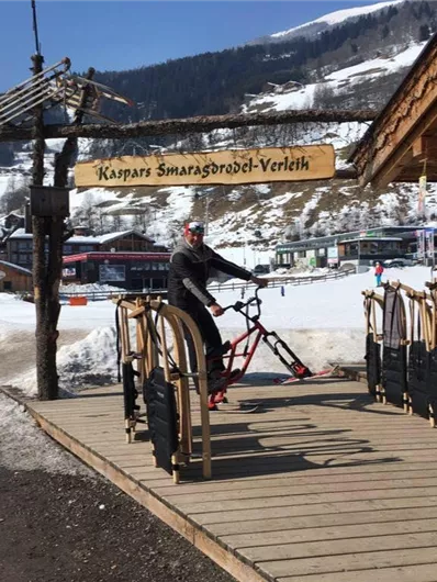 Een man rijdt op een speciale fiets voor een verhuurstalletje in het besneeuwde berglandschap. Op de achtergrond zijn hutten en besneeuwde bergen te zien.