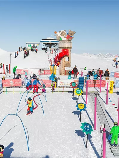 Ein lebhaftes Skigebiet mit vielen Kindern, die auf dem Schnee spielen. Bunte Spielstationen und eine klare Berglandschaft im Hintergrund.