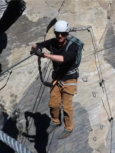 Ein Kletterer befestigt sich an einer Felswand. Er trägt einen Helm und Kletterausrüstung und ist von einer beeindruckenden Naturkulisse umgeben.