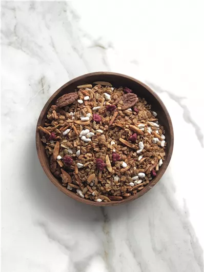 Een kom met knapperige muesli, die noten, gedroogd fruit en verschillende granen bevat. De mix heeft een aantrekkelijke, gouden kleur en is geplaatst op een gemarmerd oppervlak.