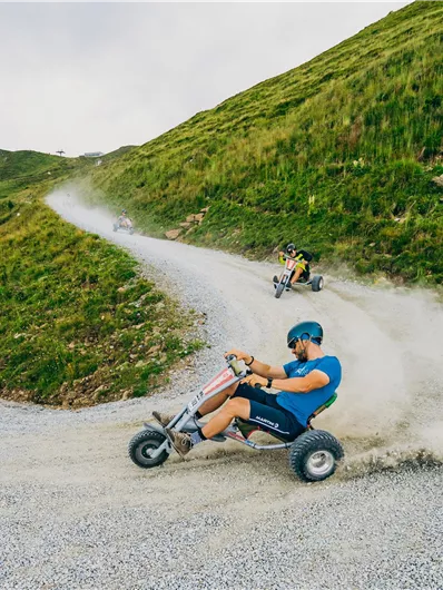 A group of people is riding tricycles down a winding path on the hill. Dust is swirling up as they have fun riding.