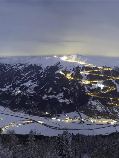 Eine winterliche Berglandschaft bei Nacht mit schneebedeckten Hügeln und beleuchteten Dörfern. Der Himmel ist düster, aber die Lichter der Stadt glänzen im Schnee.