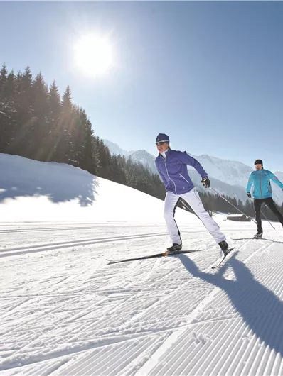 Zwei Skifahrer fahren über eine weiße Piste in der Sonne. Im Hintergrund sind schneebedeckte Berge und Bäume zu sehen.