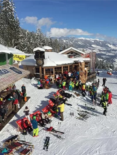 Ein belebter Skihang mit vielen Menschen, die Skifahren und sich entspannen. Im Hintergrund sind schneebedeckte Bäume und Berge zu sehen.
