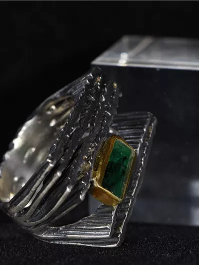 Ein eleganter Ring aus Silber mit einem grünen Stein und goldenen Akzenten. Der Ring steht neben einem klaren, quadratischen Block.