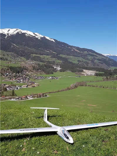 Ein Segelflugzeug auf einer grünen Wiese mit Blick auf eine hügelige Landschaft und schneebedeckte Berge im Hintergrund. Der Himmel ist klar und blau.