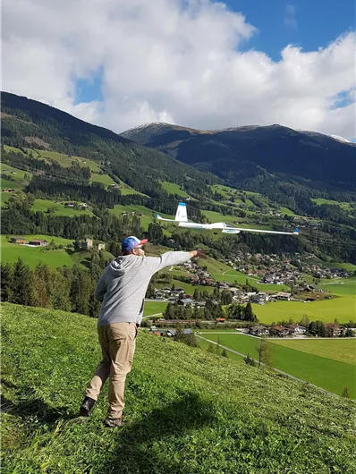 Ein Mann steht auf einer Wiese und zeigt auf eine Stadt im Tal. Die Umgebung ist grün mit Bergen im Hintergrund und einem blauen Himmel.