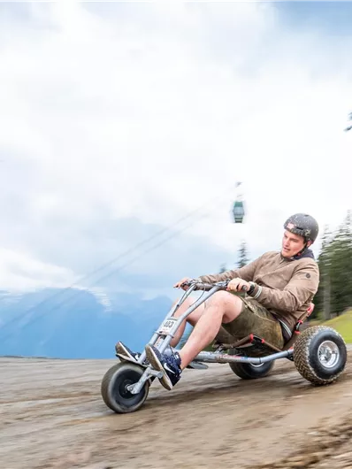 A man is riding on a trail in a mountain cart. In the background, there are trees and a cable car visible.