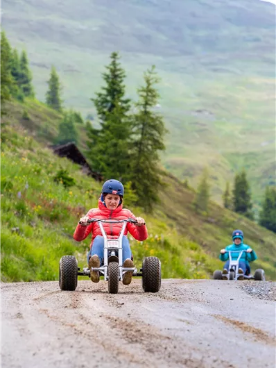 Two people are driving on a gravel path in go-karts. The surroundings are green and mountainous, ideal for adventure.