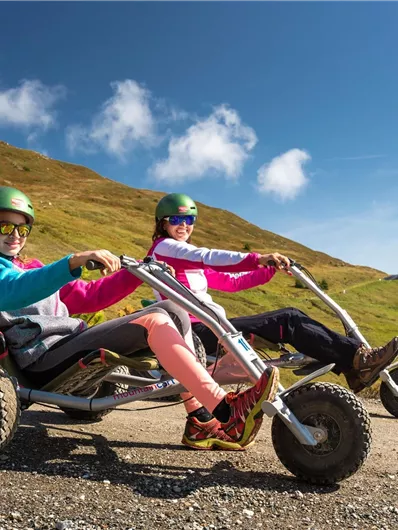 Zwei Frauen fahren auf Mountain-Carts auf einer malerischen Strecke im Freien. Die Umgebung ist von sanften Hügeln und blauem Himmel umgeben.