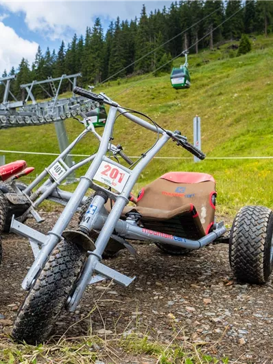 A few mountain cart vehicles are standing on a gravel path. In the background, there is a green meadow and a cable car visible.