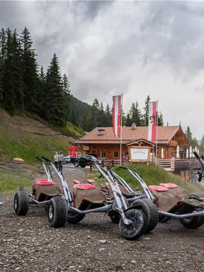 A view of a mountain hut in the background, surrounded by trees. In the foreground, several off-road vehicles are present.