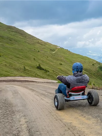 A person is riding a mountain cart down a narrow, winding path downhill. The landscape is green, and the clouds in the sky give the scene an exciting atmosphere.