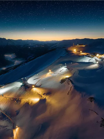 Ein atemberaubender Blick auf eine schneebedeckte Berglandschaft bei Nacht. Die Lichter der Pisten und Anlagen strahlen im Dunkeln, während der Himmel voller Sterne funkelt.