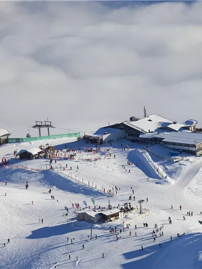 Eine schneebedeckte Berglandschaft mit vielen Skifahrern und einer Berghütte. Der Himmel ist klar und die Wolken liegen tief unterhalb der Piste.