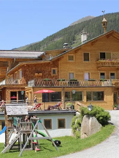 Ein gemütliches Holzhaus in den Bergen mit einer Terrasse und Tischen im Freien. Im Vordergrund gibt es einen Kinderspielplatz und eine schöne Landschaft.