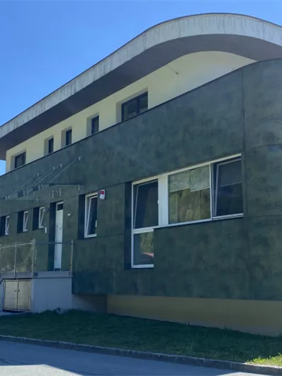 Ein modernes Gebäude mit einer ungewöhnlichen, grünen Fassade. Es hat große Fenster und eine umlaufende Fläche im Obergeschoss.