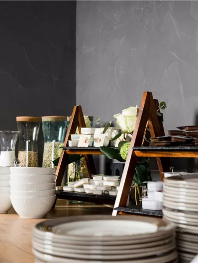 Een stijlvolle tafel met witte kommen, borden en decoratieve glazen. Op de achtergrond staan houten planken met een selectie van snacks en bloemen.