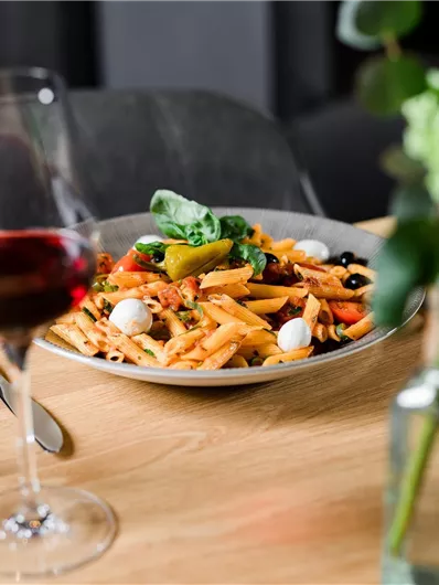 Een bord pasta met kleurrijke groenten en mozzarella, geserveerd op een houten tafel. Op de achtergrond staan een glas rode wijn en een bloemenvaas.