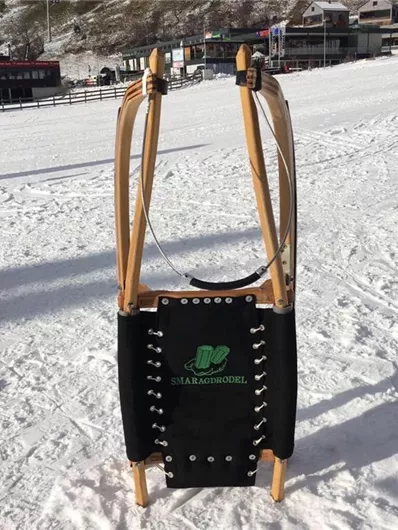 Een slee staat op de sneeuw voor een berglandschap. Op de achtergrond zijn huizen en voorzieningen zichtbaar.