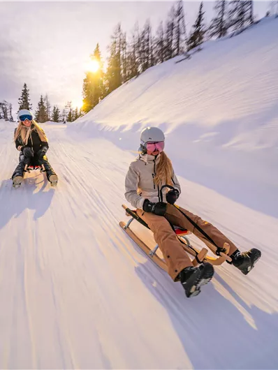 Zwei Personen rodeln auf einer verschneiten Piste. Die Sonne scheint im Hintergrund und der Himmel ist bewölkt.