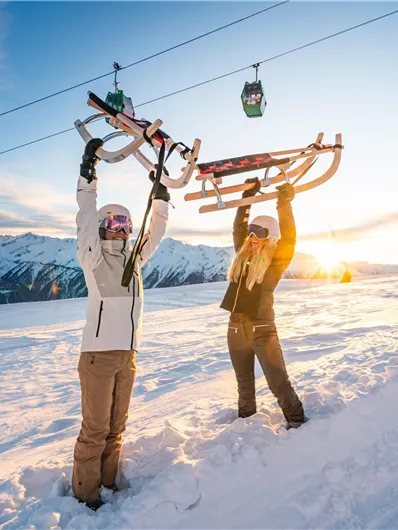 Zwei Personen stehen im Schnee und halten Schlitten in die Höhe. Im Hintergrund sieht man einen Skilift und eine beeindruckende Berglandschaft bei Sonnenuntergang.