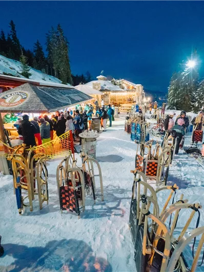 Eine alpine Skihütte bei Nacht, umgeben von schneebedeckten Bäumen. Viele Menschen genießen die winterliche Atmosphäre und die Geselligkeit im Freien.