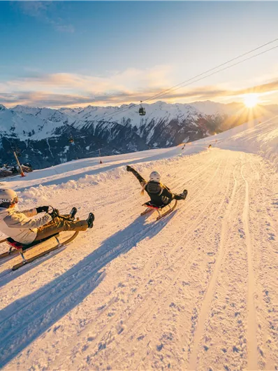 Zwei Schlittenfahrer auf einer schneebedeckten Piste mit herrlichem Bergpanorama. Die Sonne geht langsam hinter den Bergen unter.