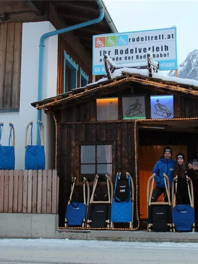 Een houten gebouw met een ingang, waar twee personen voor staan. Naast de ingang zijn sleeën en blauwe rugzakken tentoongesteld, terwijl op de achtergrond besneeuwde bergen te zien zijn.